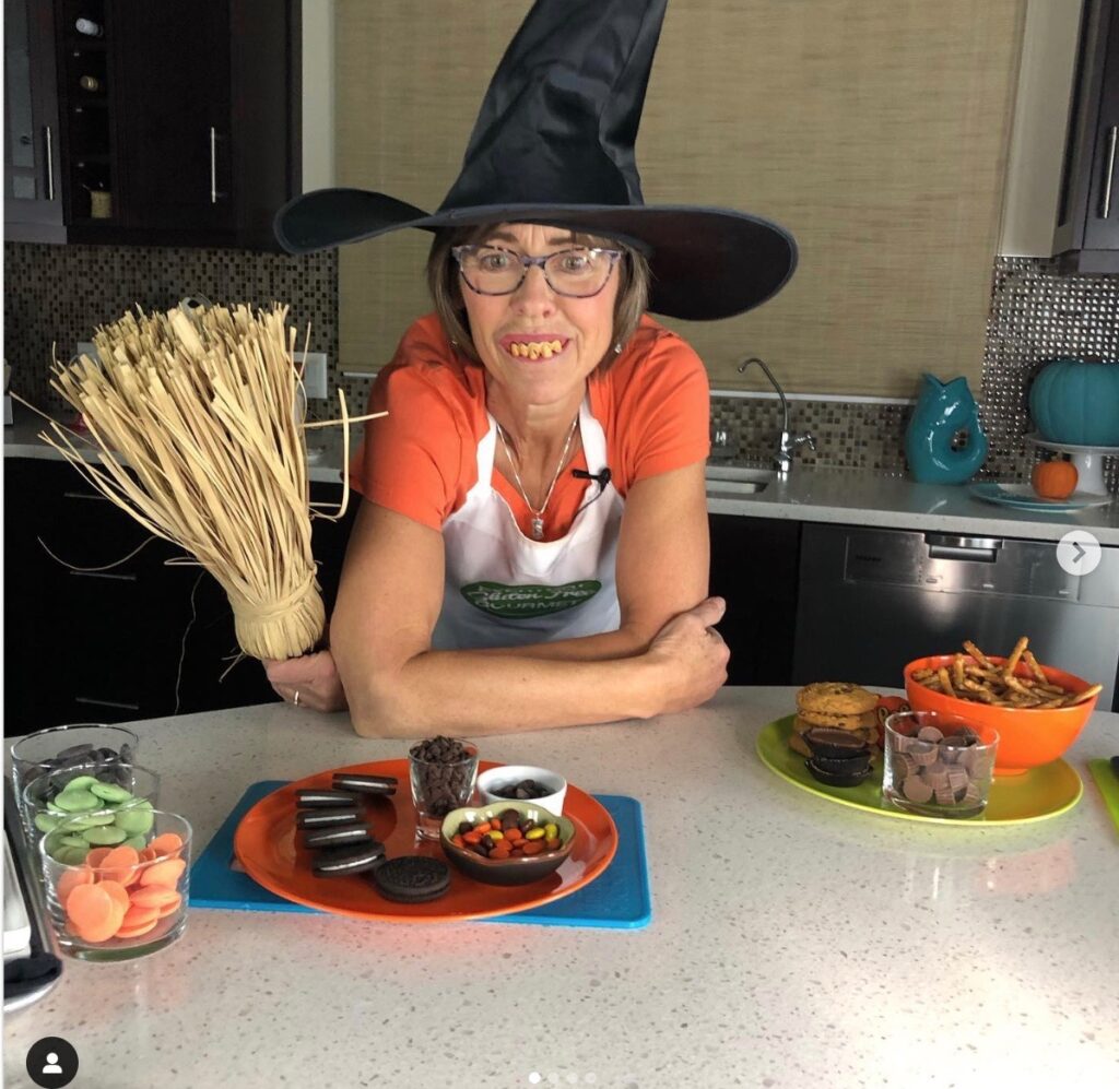 Cinde in her kitchen wearing an orange shirt, witch hat and crooked teeth holding a broom. Bowls of Halloween candy in front of her.