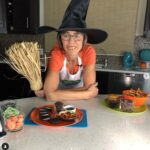 Cinde in her kitchen wearing an orange shirt, witch hat and crooked teeth holding a broom. Bowls of Halloween candy in front of her.