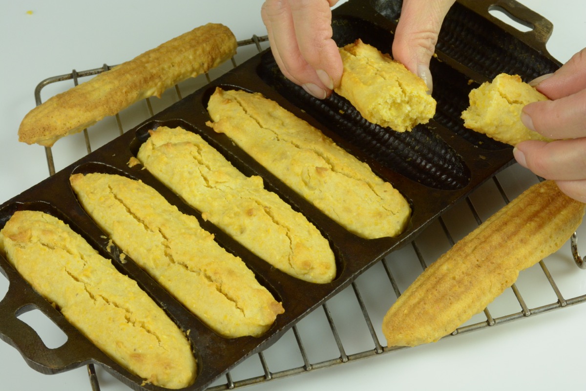 A cast iron corn stick mold with cooked cornbread and a hand holding one with a bite out of it.