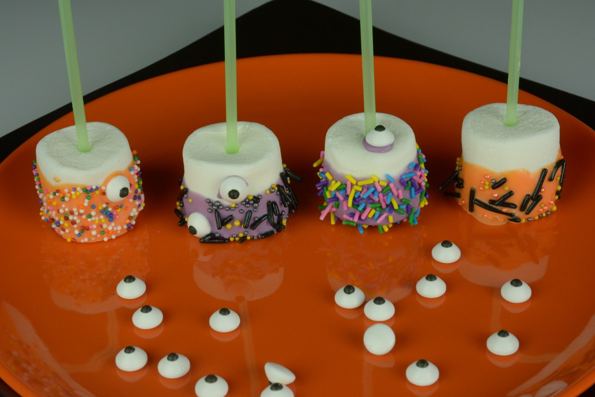 On orange plate with 4 marshmallows that have been dipped in coloured chocolate then sprinkles and a few googly eyes.