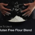 Two floured hands, wiping flour on a black apron from a bowl of flour in front of them.