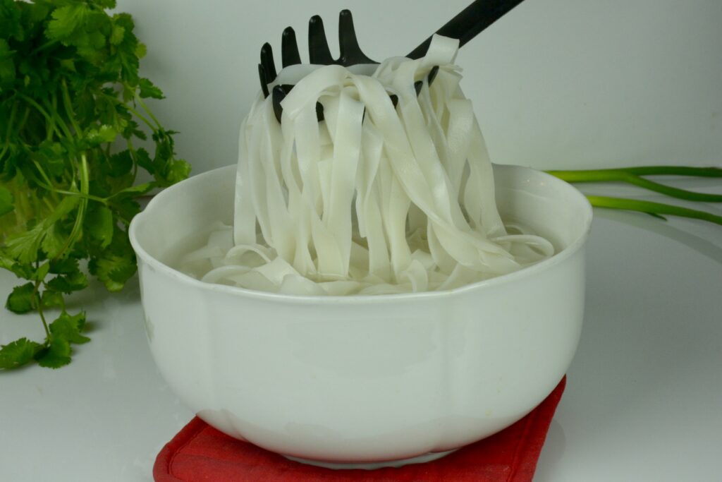 A pasta stirrer lifting softened rice noodles out of a large, white bowl.
