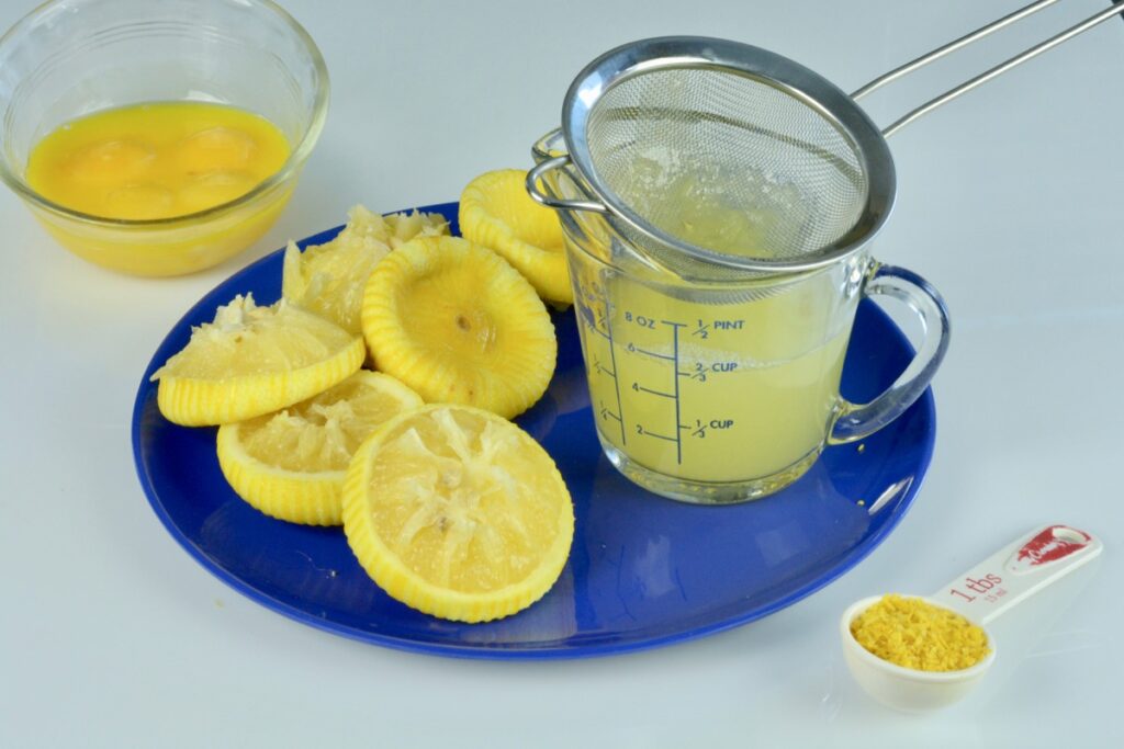 A bowl of egg yolks, a cup of lemon juice and a plate with all the squeezed lemon halves.