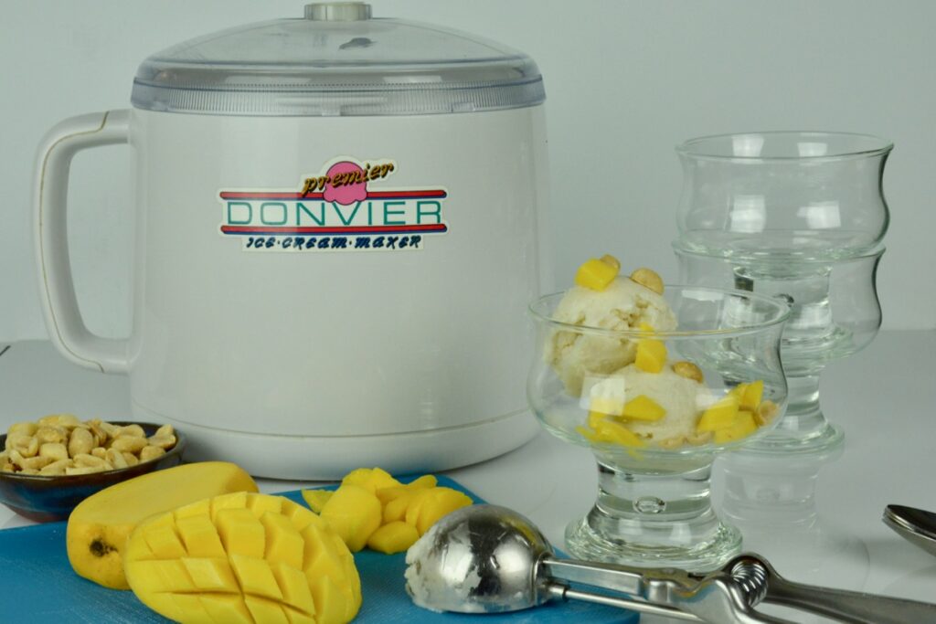 A Donvier ice cream maker surrounded by cut mango, a dish of peanuts and a serving of coconut ice cream with toppings.