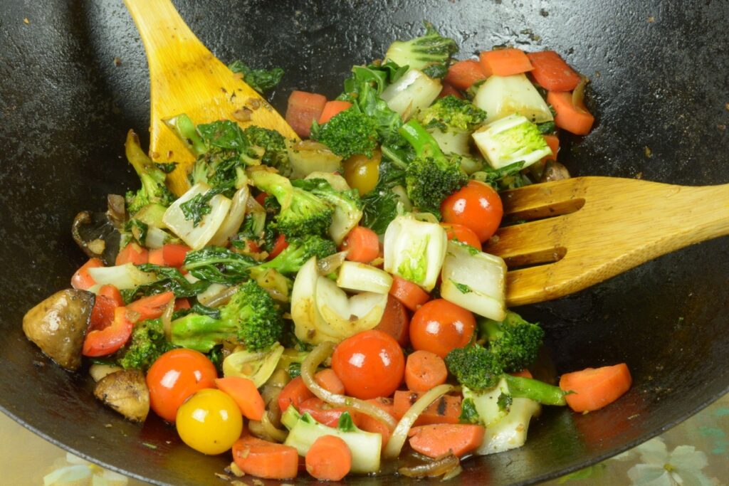 A wok full of colourful vegetables and two wooden spoons stirring them.