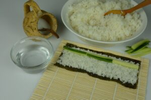 A bamboo mat with a half sheet of nori, covered in rice an topped with a long row of cucumber, ready to be rolled into hosomaki.