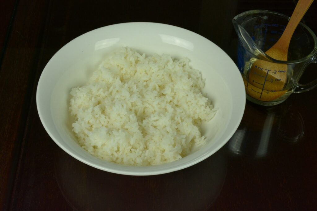 A white bowl filled with cooked sushi rice and a cup with the seasoned rice vinegar ready to mix.