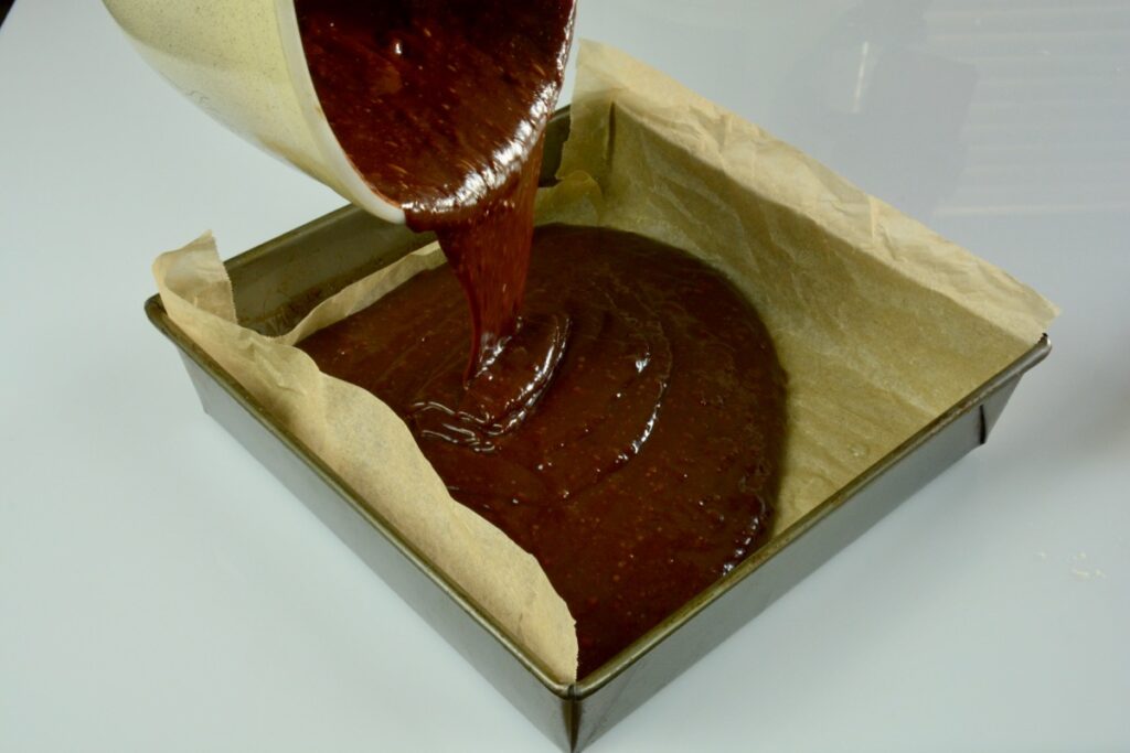 Brownie batter being poured into a baking pan lined with parchment.