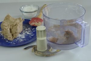 A food processor beside a blue plate with the gluten free pasta dough on it and a small bowl of tapioca starch.