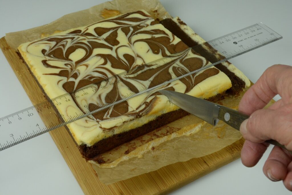 A pan of Chocolate Cheesecake Brownies on a cutting board with a ruler laying across the top and a knife marking them at 1-inch intervals.