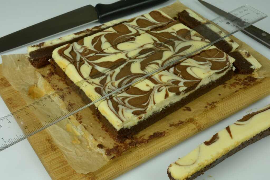 A pan of Chocolate Cheesecake Brownies on a cutting board with the edge cut off for a perfect clean cut. A ruler is laying across the brownie ready to mark the cut lines at 1-inch intervals.