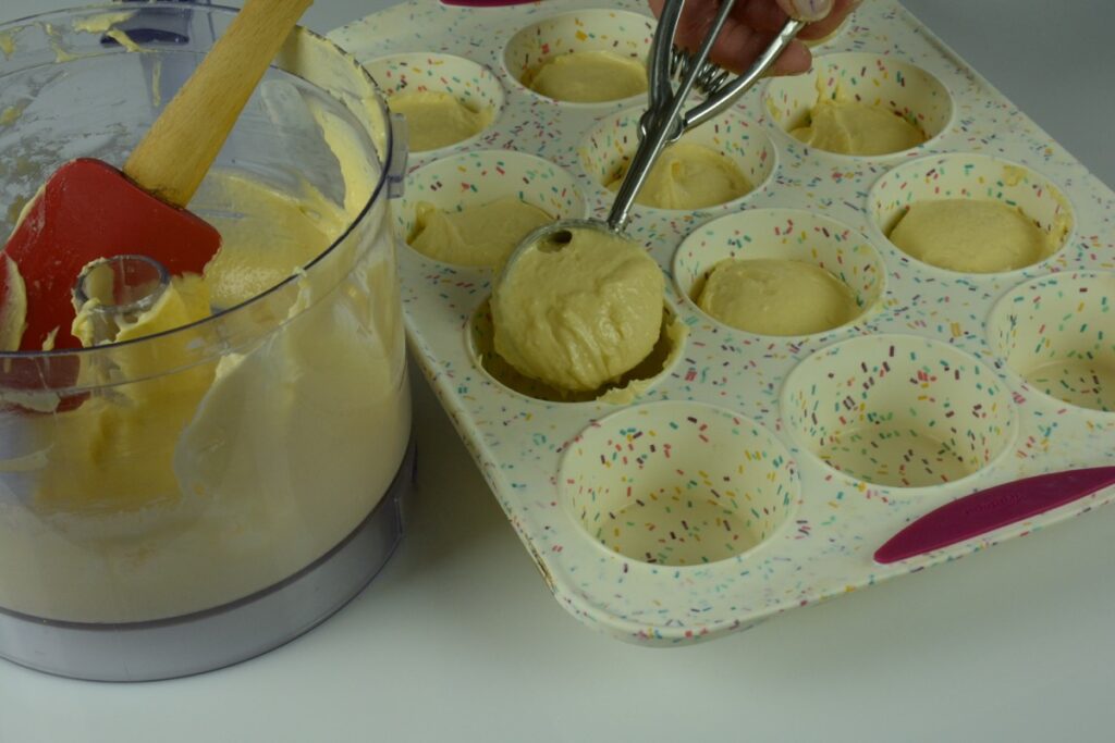 A silicone muffin pan being filled with batter for cheese buns using a metal scoop.