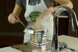Someone cranking a piece of gluten free pasta through the fettuccini setting of a hand crank pasta machine.