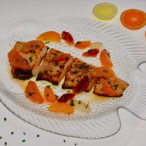 A clear plate with pieces of baked Arctic Char covered in citrus sauce with whole segments of citrus fruit and a sprinkling of chives.
