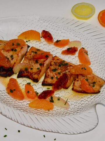 A clear plate with pieces of baked Arctic Char covered in citrus sauce with whole segments of citrus fruit and a sprinkling of chives.