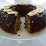 Banana Bundt Cake on a pedestal tray drizzled with Chocolate Glaze and topped with banana slices.