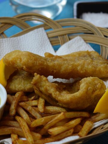 A basket full of gluten free Battered Fish and Chips with lemon wedges and a dish of tartar sauce.
