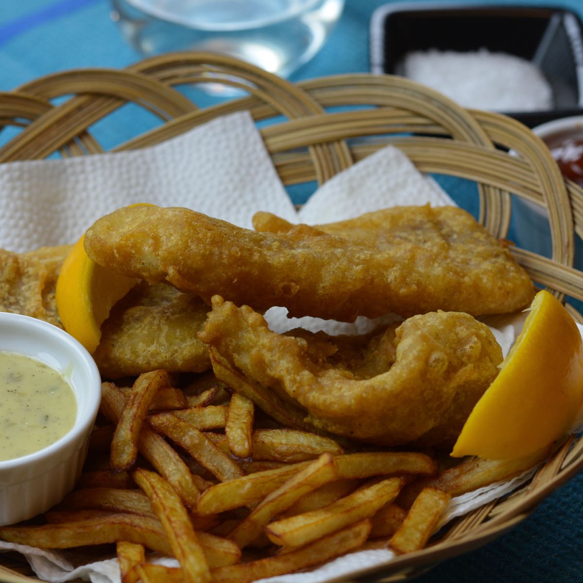 A basket full of gluten free Battered Fish and Chips with lemon wedges and a dish of tartar sauce.