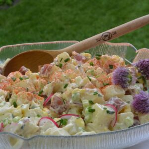 A wide bowl of Potato Salad with radish slices, snipped chives, a sprinkling of paprika and a few chive blossoms. Sitting on grass with a wooden spoon in it.