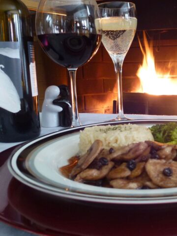 A plate of Chicken Porto with rice pilaf and broccoli on a table set in front of a fireplace.