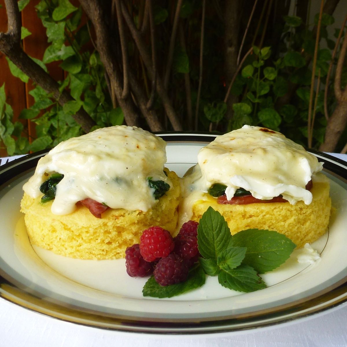 A white plate with gluten free cornbread rounds topped with ham, spinach, eggs and Mornay sauce, Eggs Florentine.