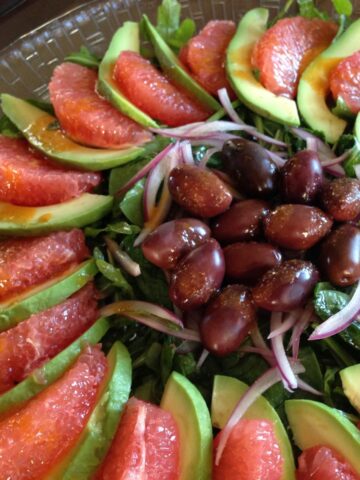 A composed Grapefruit and Avocado Salad on a bed of arugula with red onion slivers and black olives.