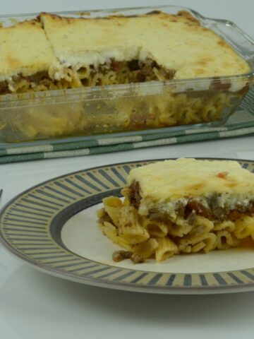 A casserole of Greek Pastitsio, a layered dish of pasta, meat sauce and white sauce; with a single piece on a plate in front of it.