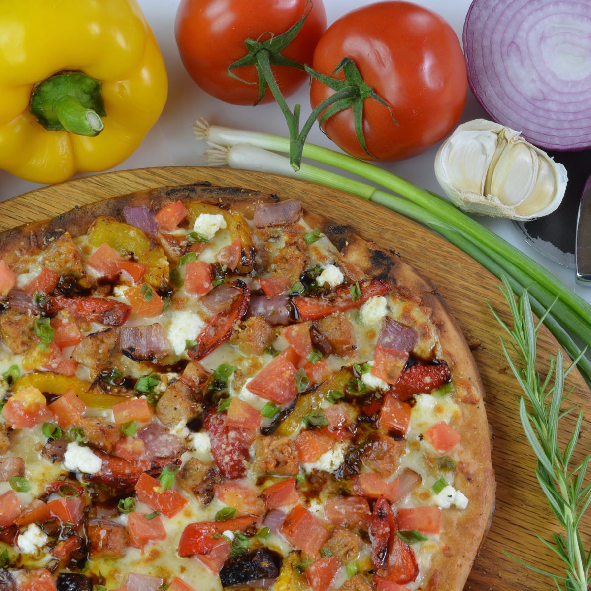 A cooked gluten free Grilled Sausage and Vegetable Pizza surrounded by peppers, tomatoes and onion.