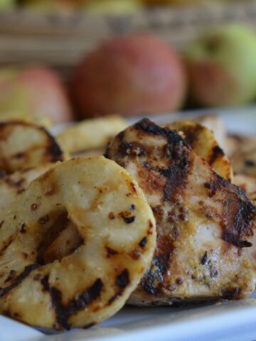 A platter of Grilled Mustard Chicken with Apple Rings with a row of red apples in the background.