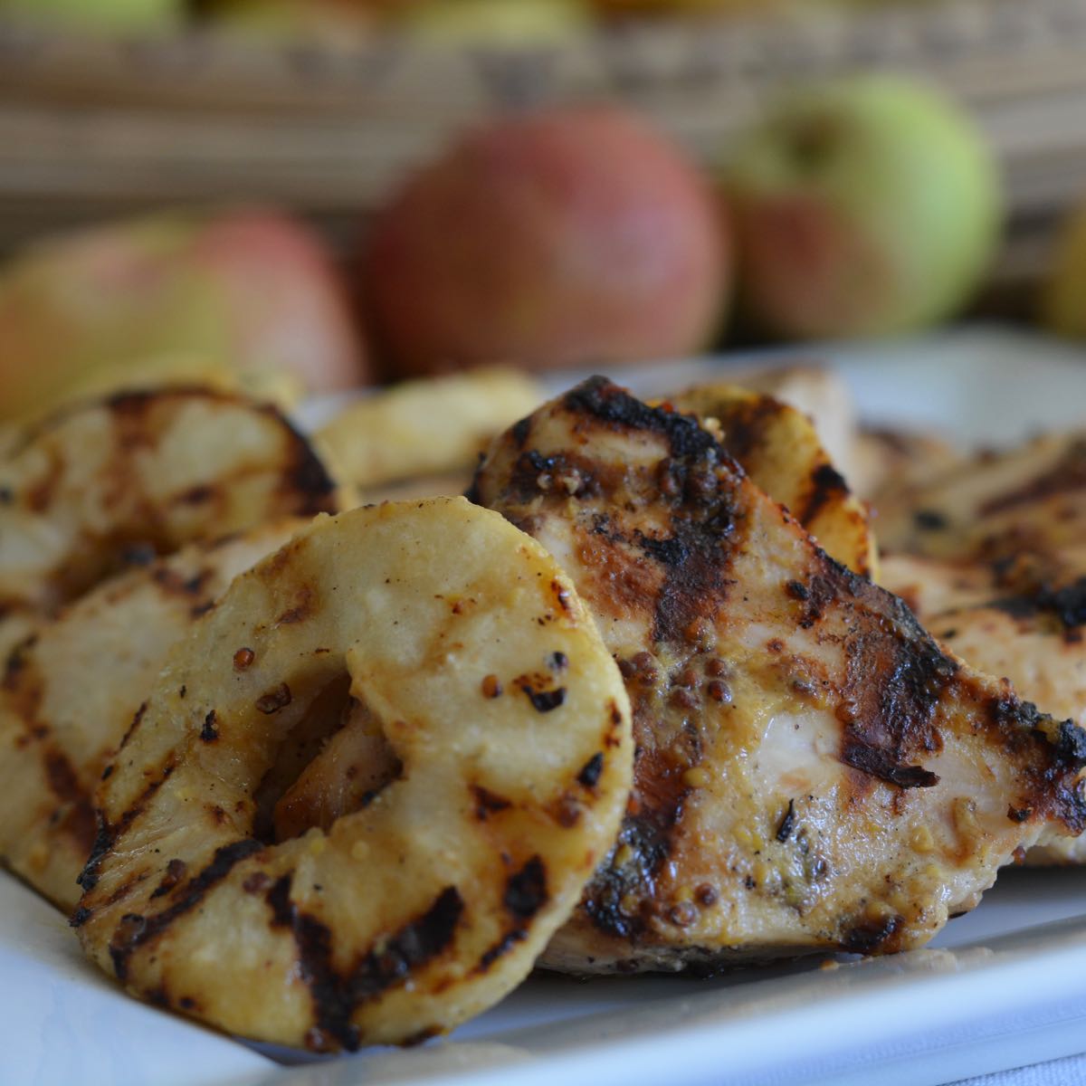 A platter of Grilled Mustard Chicken with Apple Rings with a row of red apples in the background.