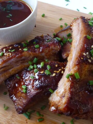 A cutting board with hoisin glazed pork ribs sprinkled with sesame seeds and chopped green onion with extra sauce on the side.