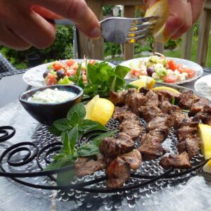 Lamb souvlaki on circular skewers served with tadziki and lemon wedges, surrounded by individual plates of Greek salad.