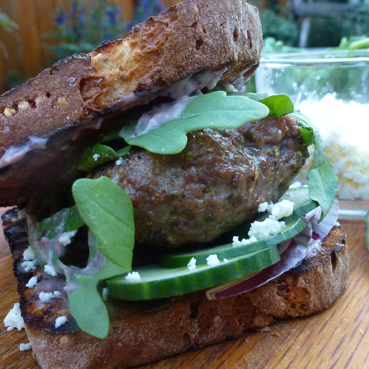 Close up of a Mediterranean Grilled Lamb Burger on toasted bread with cucumber, arugula, feta, red onion and olive mayo.