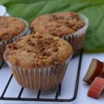 A baking rack with rhubarb streusel muffins surrounded by chopped rhubarb and a rhubarb leaf.