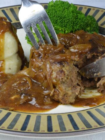 A plate with a fork and knife cutting a Salisbury Steak surrounded by mashed potatoes and Onion Gravy.
