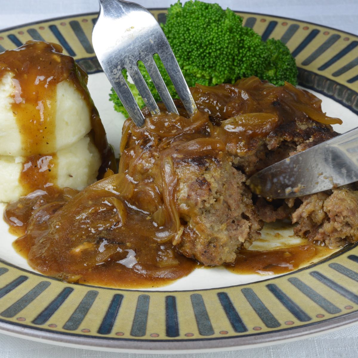 A plate with a fork and knife cutting a Salisbury Steak surrounded by mashed potatoes and Onion Gravy.
