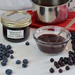 A jar of homemade Saskatoon Blueberry Barbecue Sauce surrounded by berries and the pot they were cooked in.