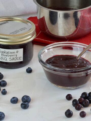 A jar of homemade Saskatoon Blueberry Barbecue Sauce surrounded by berries and the pot they were cooked in.