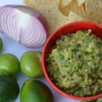 A bowl of tomatillo guacamole surrounded by limes, tomatillos, red onion and tortilla chips.