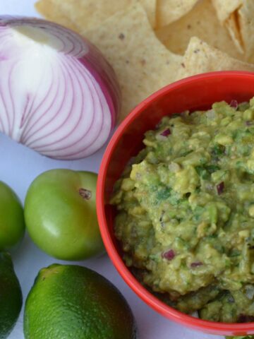 A bowl of tomatillo guacamole surrounded by limes, tomatillos, red onion and nacho chips.