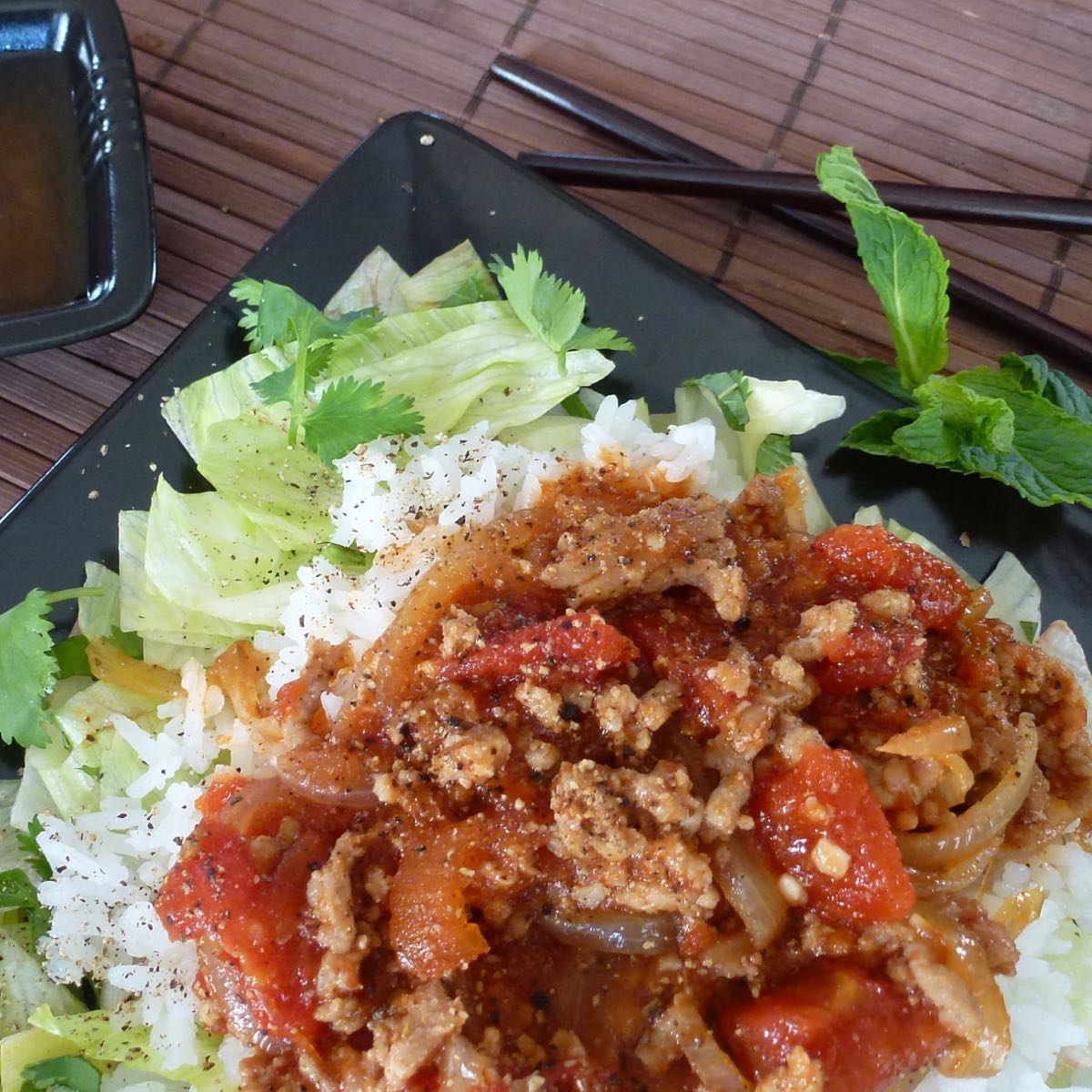 A square black plate with a bottom layer of lettuce and fresh herbs, covered with warm rice and topped with Vietnamese Pork in Tomato Sauce. A small black dish with nuoc cham beside it.