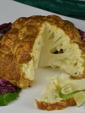 A whole roasted cauliflower on a platter lined with lettuce leaves and one wedge cut out topped with a twist of lime.