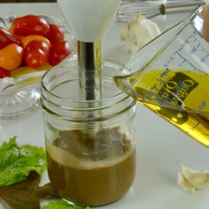 Olive oil being poured into a Mason jar with a hand blender in it, surrounded by lettuce, garlic and a whisk.