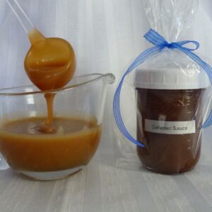 A jar of Homemade Caramel Sauce wrapped and labelled for a gift; beside a sauce boat with caramel sauce on a ladle dripping back into the dish