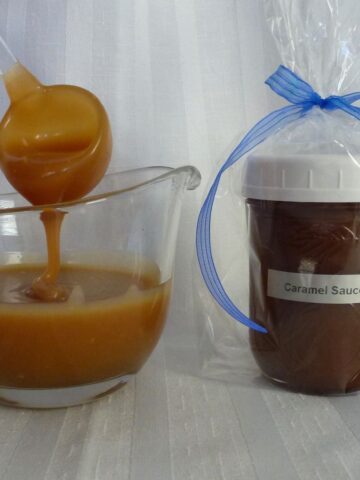 A jar of Homemade Caramel Sauce wrapped and labelled for a gift; beside a sauce boat with caramel sauce on a ladle dripping back into the dish