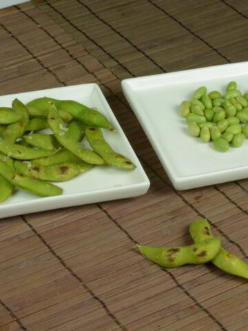A bamboo place mat with two plates; one with whole pods of edamame beans, the other with shelled beans.
