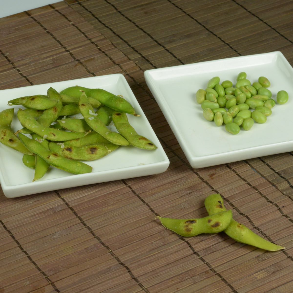 A bamboo placemat with two plates; one with whole pods of edamame beans, the other with shelled beans.