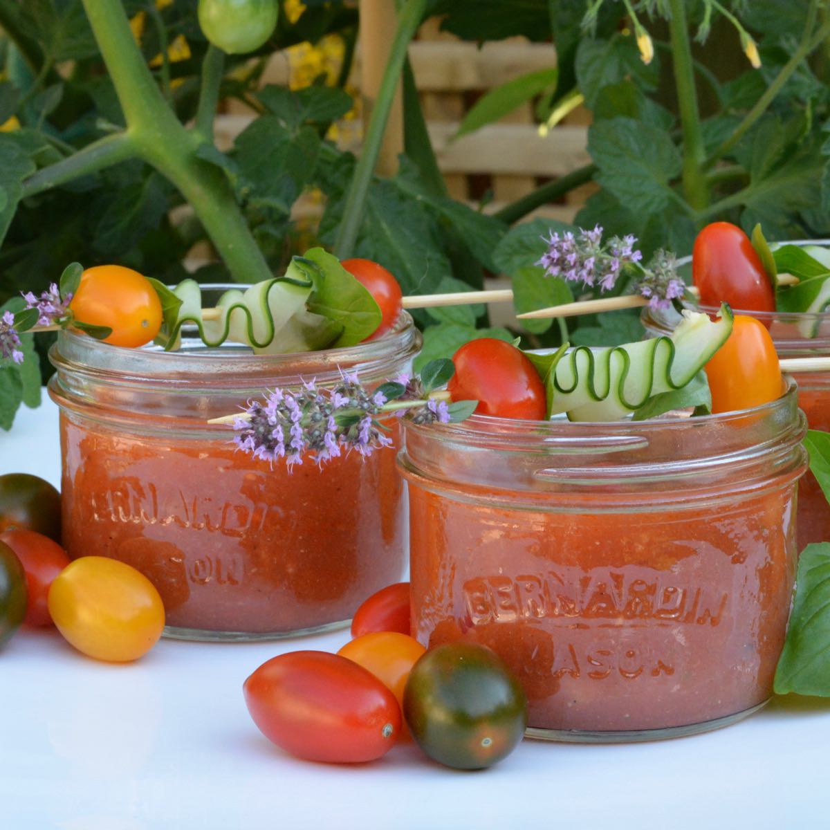 Two Mason jars filled with Gazpacho and garnished with a skewer of cucumber ribbon, fresh thyme and a cherry tomatoes.