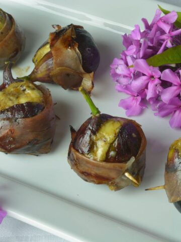 Grilled Figs Stuffed with Blue Cheese and wrapped with prosciutto on a white platter surrounded by pink flowers.