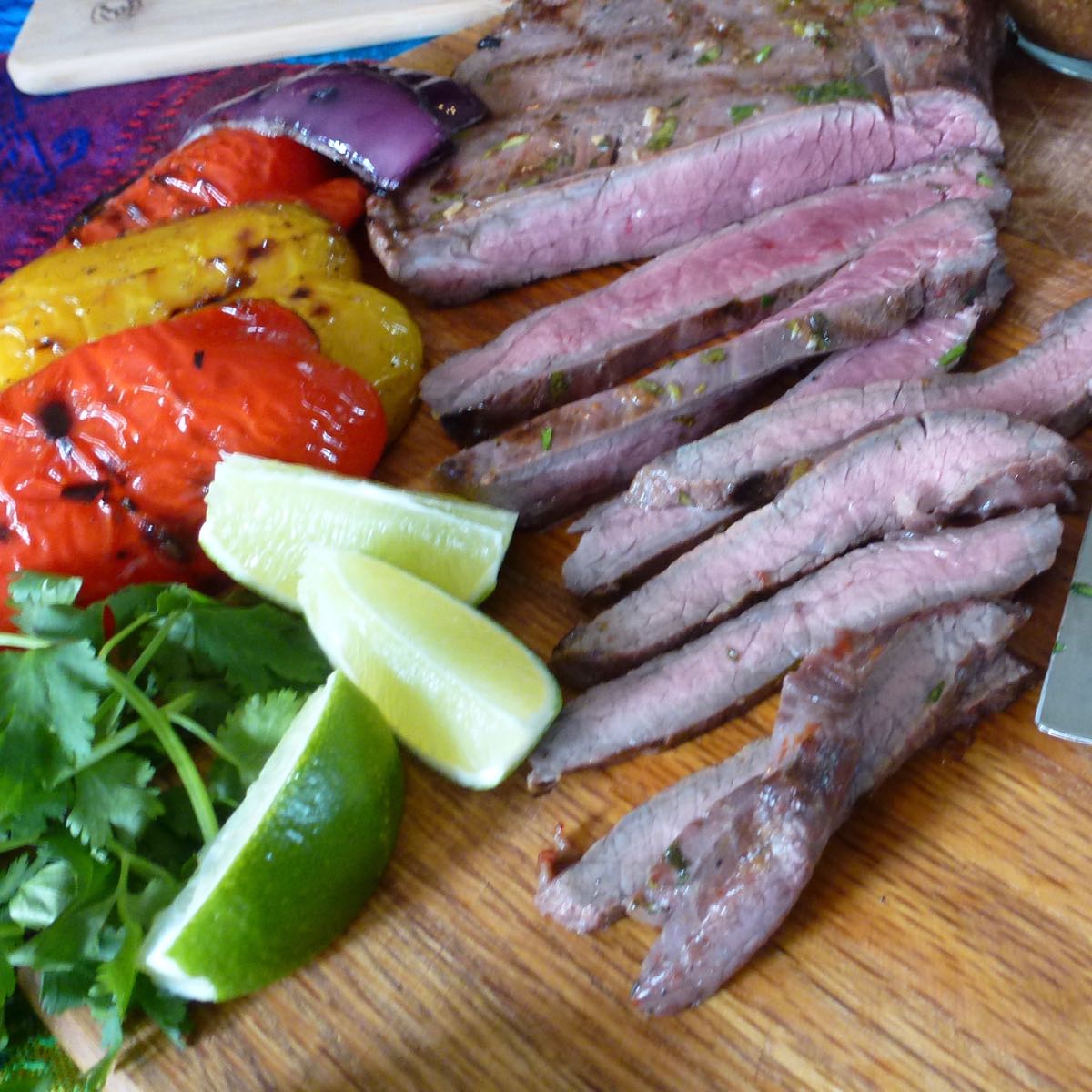 A board with slices of Grilled Flank Steak and bell peppers with lime wedges and cilantro.
